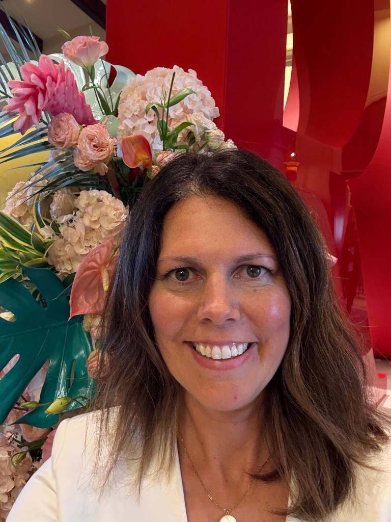Woman smiling indoors beside large floral arrangement with red sculptural backdrop