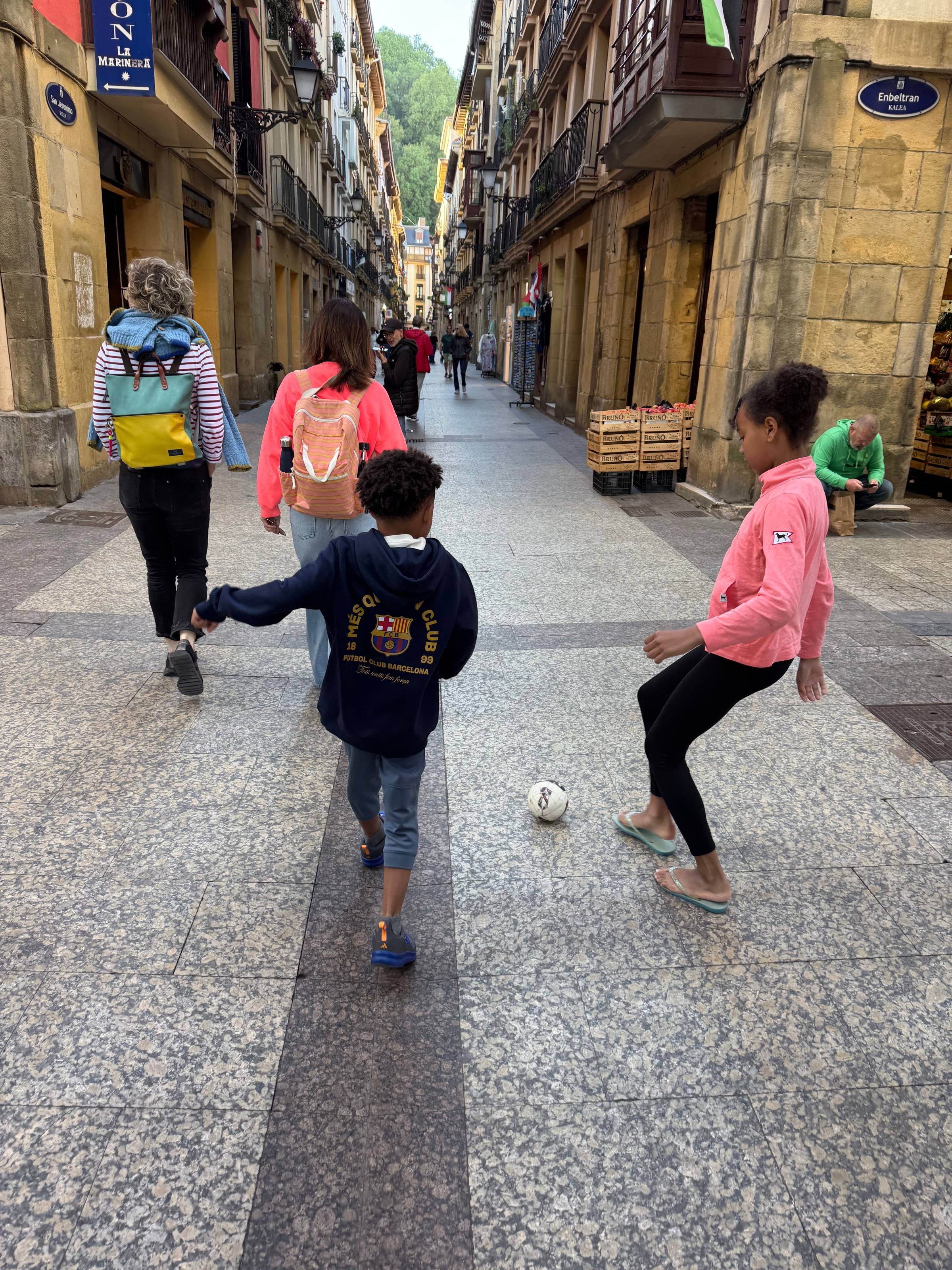 Children playing soccer on a European street while adults walk ahead, capturing real-life movement and vision in action.