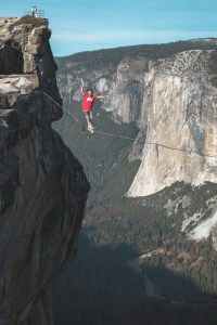 Person walking a highline between two cliffs in Yosemite, arms outstretched for balance, with El Capitan and a forested valley in the background
