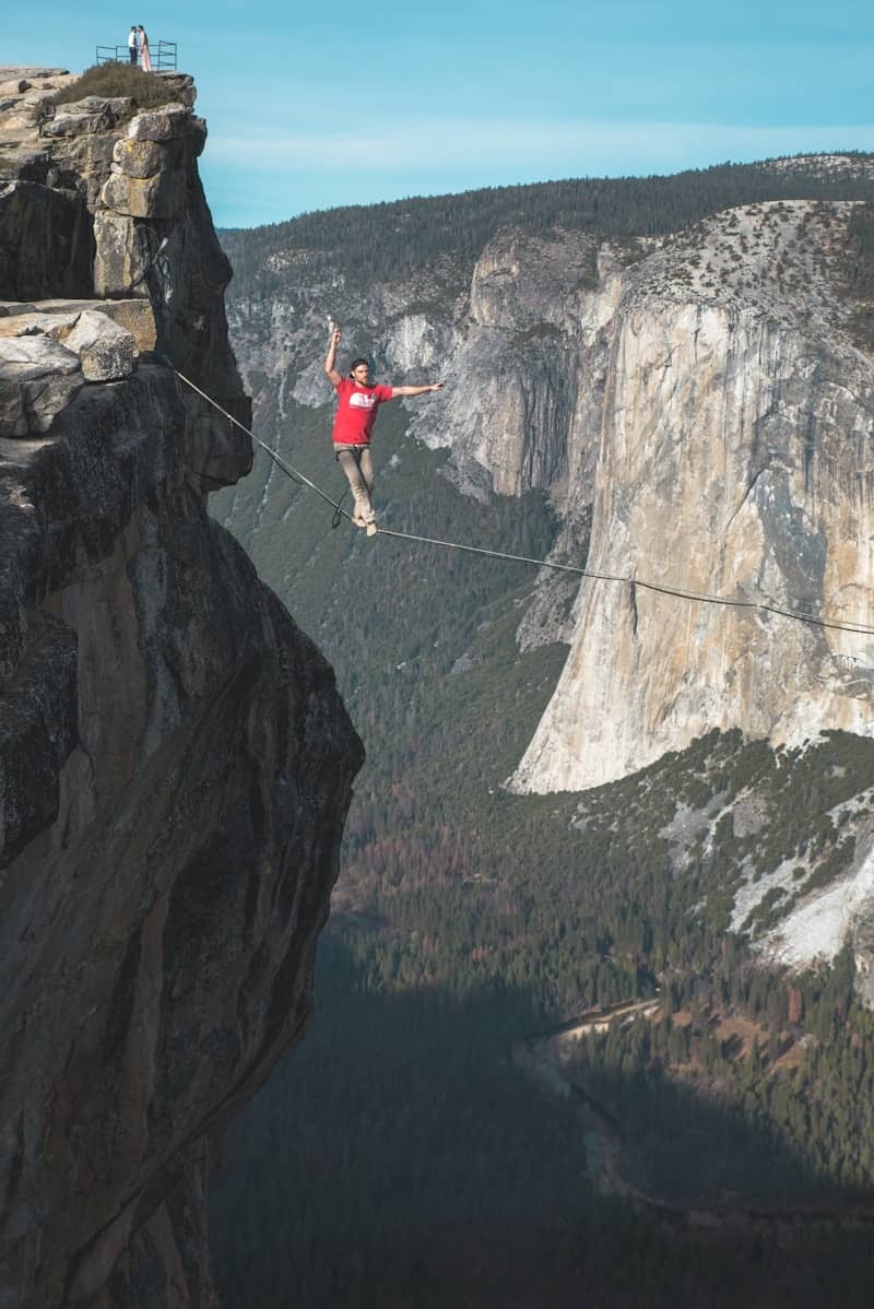 Person walking a highline between two cliffs in Yosemite, arms outstretched for balance, with El Capitan and a forested valley in the background